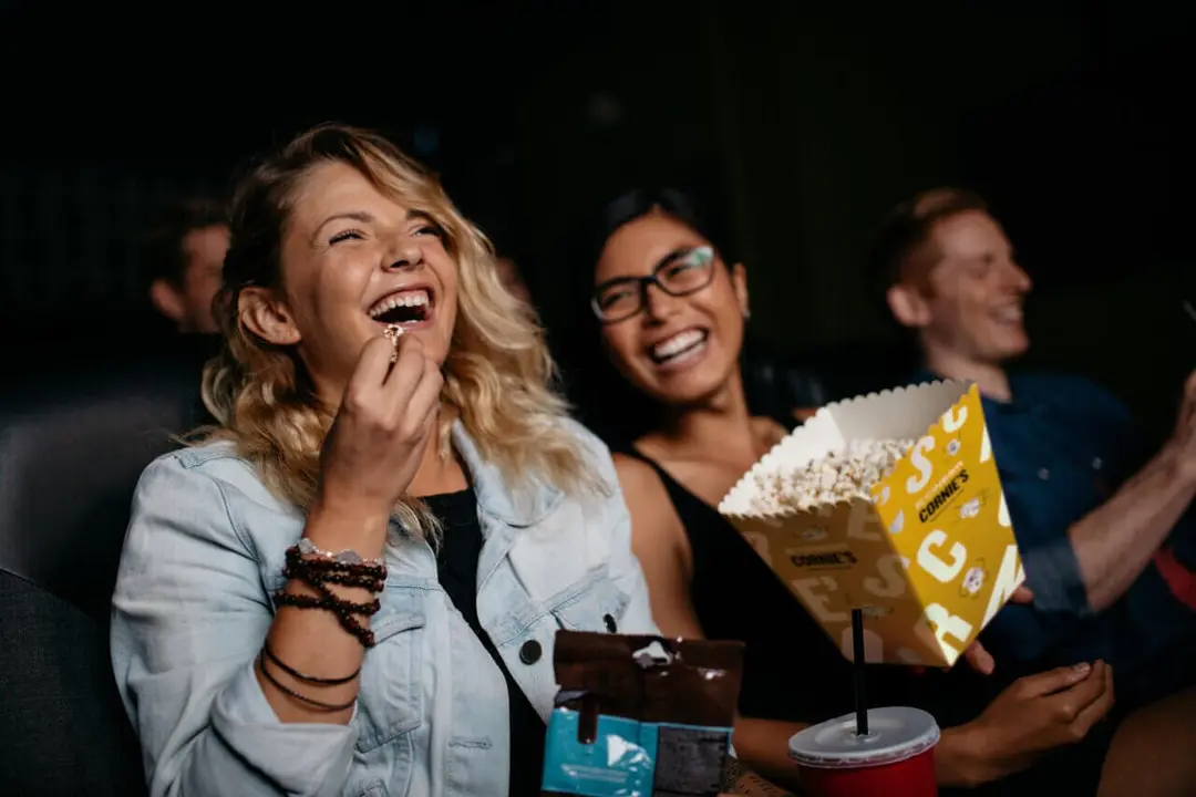 Young woman with friends watching movie in cinema and laughing. Group of people in theater with popcorns and drinks.