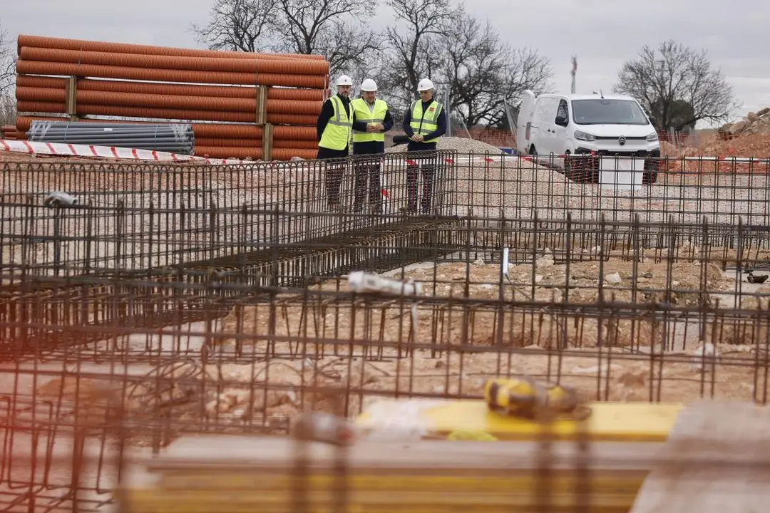 Tres trabajadores revisan los planos en un sitio de construcci&oacute;n. Se observan materiales apilados y estructuras de acero.