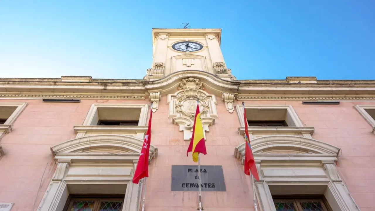 Vista de la fachada del Ayuntamiento en Alcal&aacute; de Henares, destacando su reloj y banderas.
